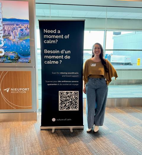 Woman standing beside Culture of Calm banner with QR code at Billy Bishop Airport promoting sound based wellness support for passengers during Autism Awareness Month